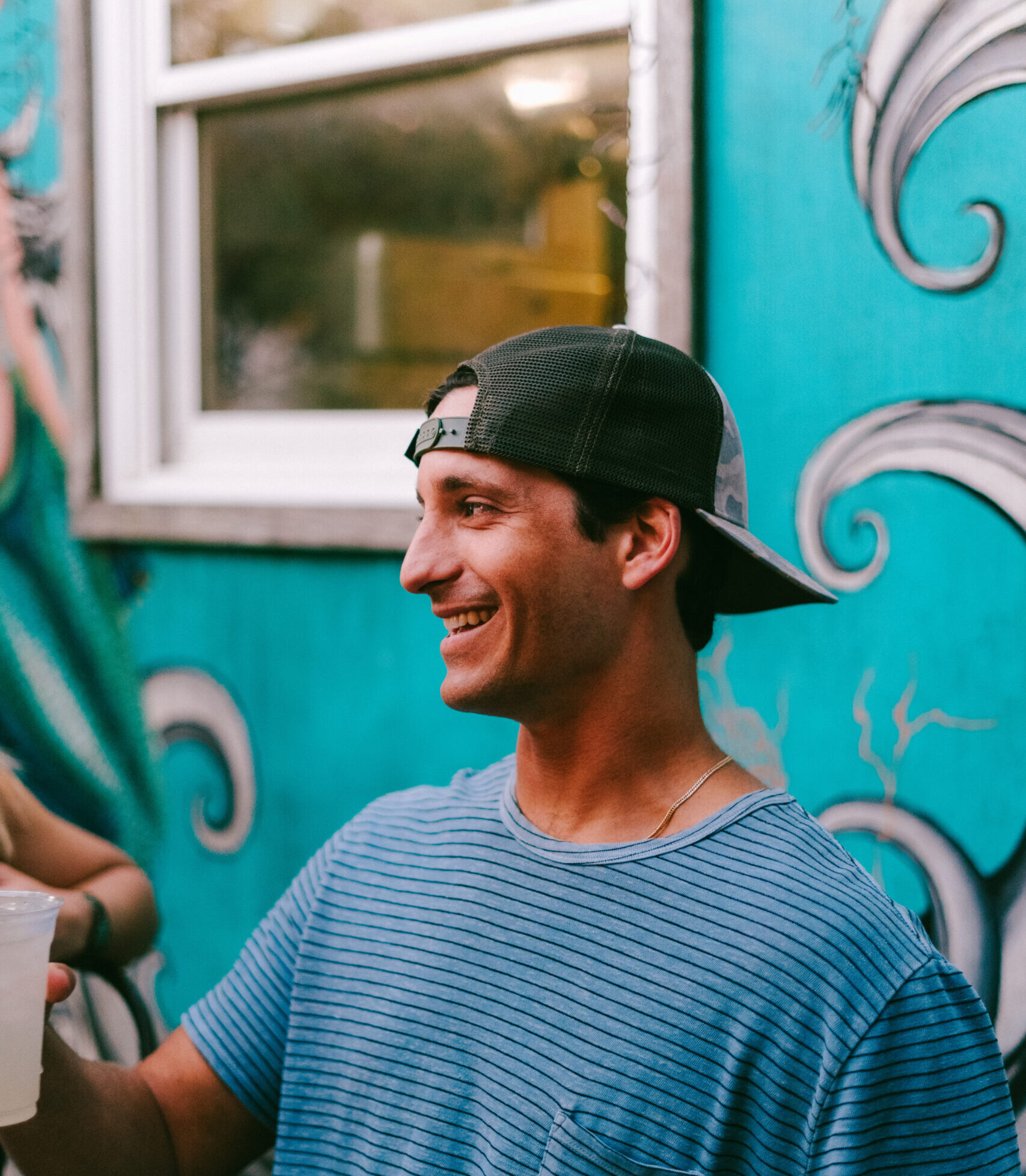 A man wearing a striped blue shirt and a backward baseball cap smiles while holding a drink in an outdoor setting. Behind him is a teal wall with artistic swirling designs, adding a lively, creative atmosphere. The scene conveys a casual and joyful moment.